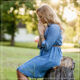 White woman with long blonde hair and denim dress sitting on a tree stump in a park praying with filtered light through trees