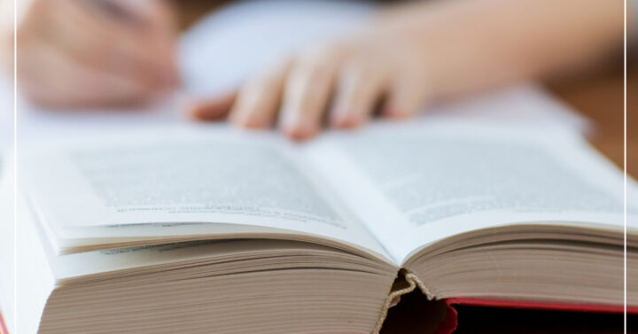 Young woman with an open hardcover book on a table writing in a notebook