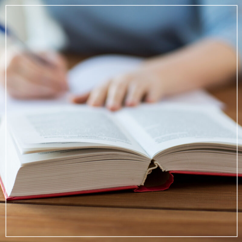 Young woman with an open hardcover book on a table writing in a notebook